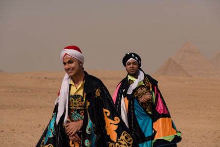 Two proud young men in vibrant Egyptian costume and turban posing together before iconic Giza pyramids in hazy desert ideal for travel and cultural