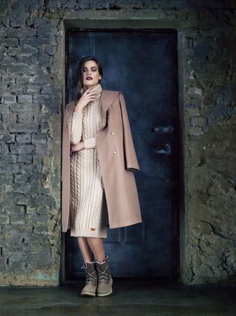 Elegant fashion model wearing long mocha coat, long beige knitted dress and boots posing before a door and an old brick wall.の写真素材