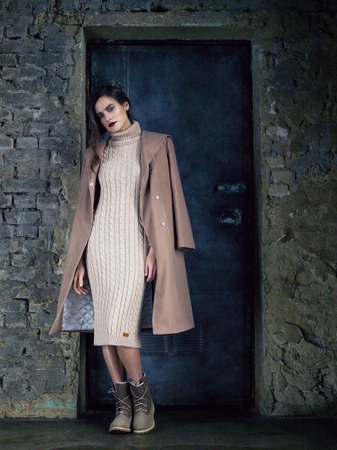 Elegant fashion model wearing long mocha coat, long beige knitted dress and boots posing before a door and an old brick wall.の写真素材