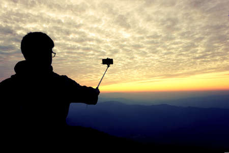 silhouette of a man taking a picture with a selfie stick on top of mountain before a sky sunset backgroundの写真素材
