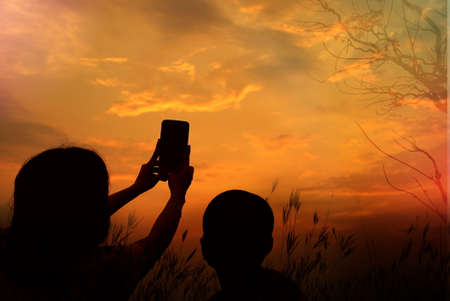 Mother and sun silhouettes taking picture by cellphone at sunset with blur nature bokeh backgroundの写真素材