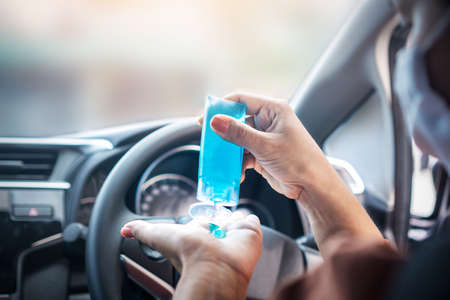 Asian woman sitting in a car applying hand sanitizer alcohol gel to clean her hand and wearing protective mask before driving during covid-19 pandemic, new normal lifestyle concept backgroundの写真素材