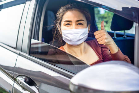 happy Asian woman hand thump up and smiling in a car wearing protective mask before driving , covid-19 pandemic, new normal lifestyle concept backgroundの写真素材