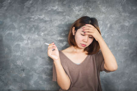 depressed Asian woman smoking cigarette with unhappy face sitting alone next to the wallの写真素材