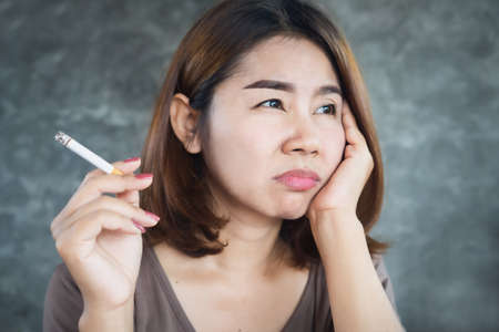 depressed Asian woman smoking cigarette with unhappy face sitting alone next to the wallの写真素材