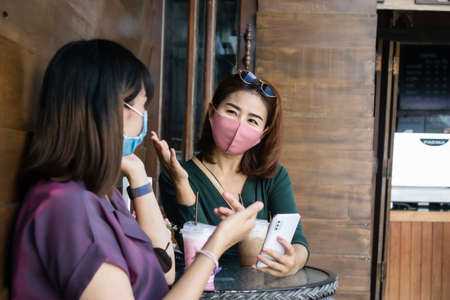 two Asian female wearing protective mask talking and drinking coffee together outdoor at a cafeの写真素材