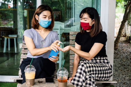 two Asian females friend wearing protective mask cleaning their hand  with alcohol gel at coffee shop, new normal conceptの写真素材