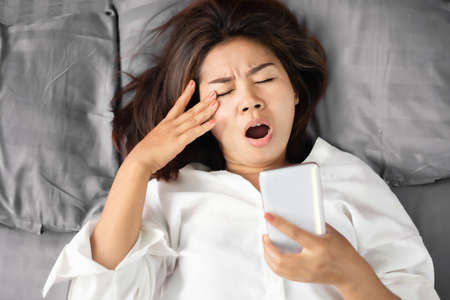 tired Asian woman yawning in bed hand holding smart phone and rubbing her eyes , social addiction conceptの写真素材