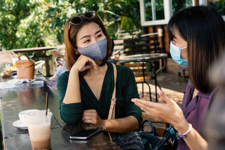 two Asian female customer wearing protective mask talking at the cafe and drinking coffee together, new normal and social distancing lifestyle conceptの写真素材