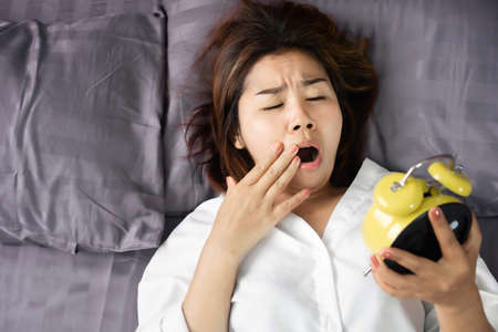 tired and sleepy Asian woman yawning in bed hand checking alarm clock lazy to wake upの写真素材