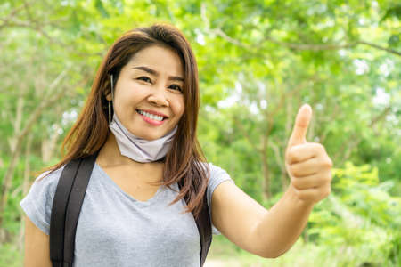 Portrait of young Asian tourism woman wearing face mask traveling alone outdoors  showing thump up hand, new normal during covid-19 conceptの写真素材
