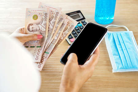 Woman hand holding smart phone and Thai banknotes 5,000 baht ,concept of covid-19 debt help or cash subsidy from governmentの写真素材