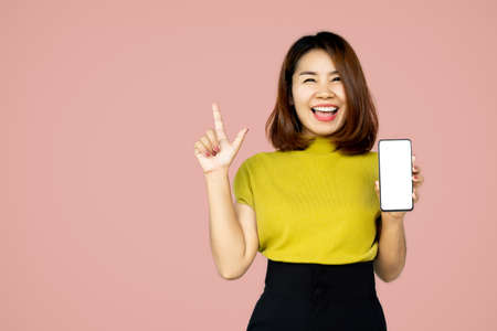 cheerful happy Asian woman hand showing smart phone and finger pointing up standing over pink backgroundの写真素材