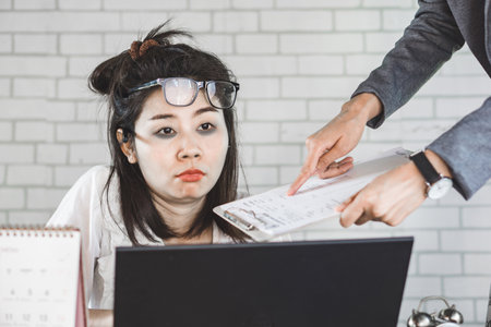 sleepless Asian woman tired and sleepy sitting at desk lazy to work with boss complaining in backgroundの写真素材