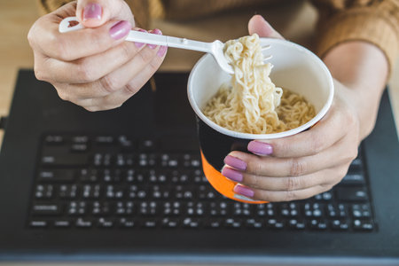 busy business woman hand eating junk food noodles cup and working on laptop at office deskの写真素材