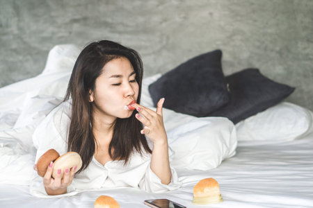 unhealthy Asian woman lying down on bed enjoy eating dessert donuts, cakeの写真素材