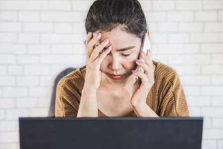 sad and depressed Asian woman talking on phone sitting at work placeの写真素材