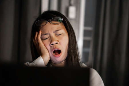 tired Asian female working late at night yawning in front of computer laptopの写真素材