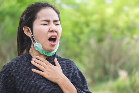 unwell Asian woman wearing mask having flu and cold sneezing outdoorsの写真素材