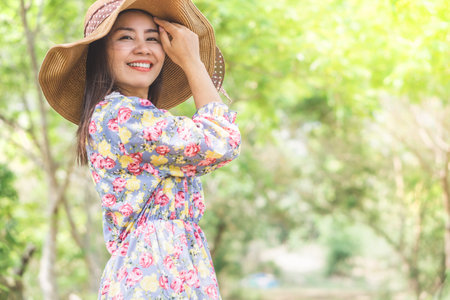 beautiful Asian woman wearing nice dress and a hat smiling at a camera with happy face relaxing outdoor in nature parkの写真素材