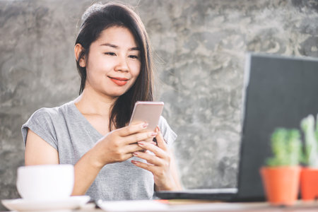 Asian female freelancer working from home office hand using smart phone with notebook on desk