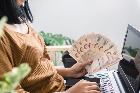 Asian woman hand holding Thai baht using laptop make money online from homeの写真素材