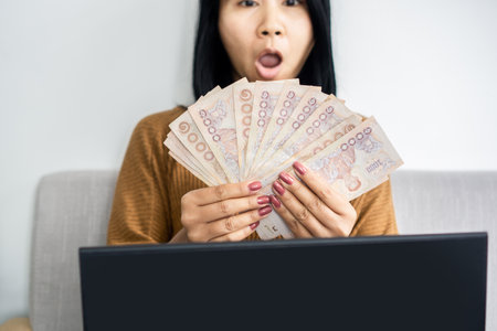 excited Asian woman showing her earning Thai baht money from working online at homeの写真素材