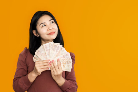 Asian woman hand holding Thai banknotes with happy face smiling over yellow background dreaming of spending moneyの写真素材