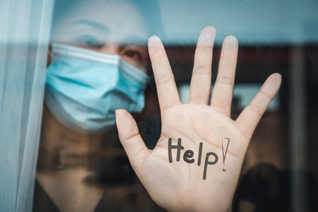 closeup woman with protective mask standing at windows show her hand with word help self-quarantine...