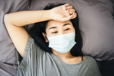 stressed Asian woman with protective face mask lying down in bed thinking or worry of sicknessの写真素材