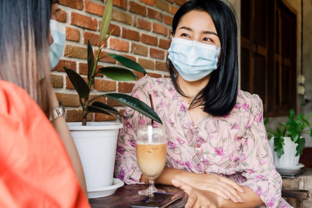 two Asian female customer wearing protective mask talking  and drinking coffee together in cafe, new normal and social distancing lifestyle conceptの写真素材