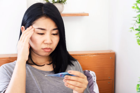 sick Asian woman having headache from fever hand holding thermometer checking high temperature