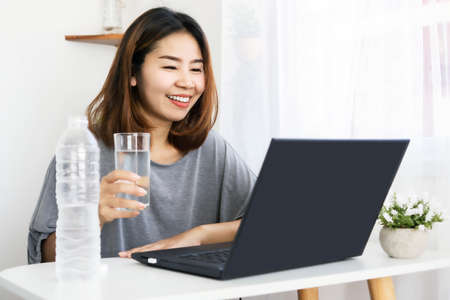 Asian woman drinking fresh water at work place sitting at office hand typing on laptop and holding glass of waterの写真素材