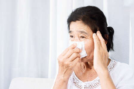 unwell old Asian woman having flu fever,  and headache hand holding paper tissue covering her runny nose and sneezingの写真素材