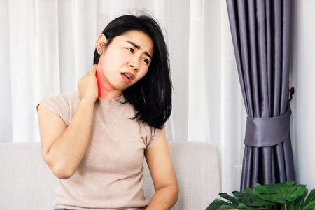 unwell Asian woman suffering from neck pain sitting on sofa hand massage her painful neck