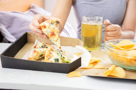 closeup woman hand eating pizza and potato chips in bed another hand holding glass of beerの写真素材
