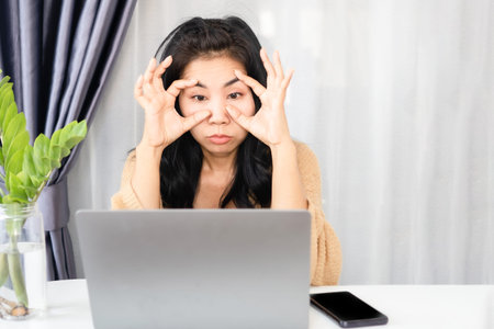 Asian businesswoman using her hand forcing her eyes open and watching laptop screen feeling tired...
