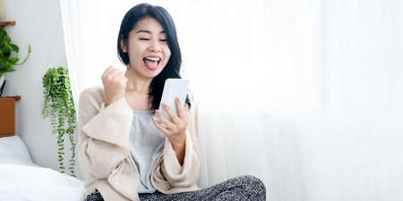 cheerful Asian woman  excited to gets good news hand holding mobile phone happy and laughing in bedの写真素材