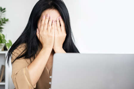 Asian businesswoman covering face in front of computer at office desk feeling tired, depressed from overworkedの写真素材