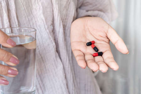 woman hand taking antibiotic pills red and black capsules with glass of waterの写真素材