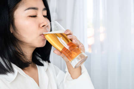 alcoholic Asian woman drinking beer hand holding a glass of beers standing next to the windowsの写真素材