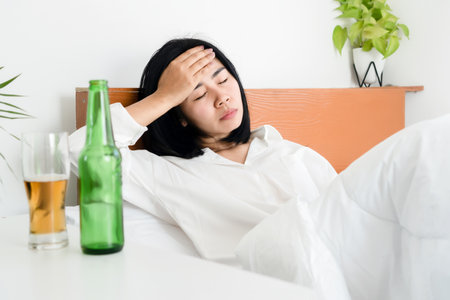 Asian woman hangover and having a headache after drinking beer wakeup in morning lying down in bedの写真素材