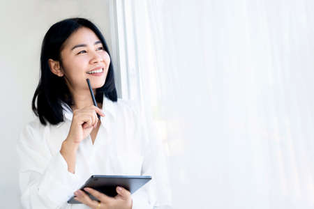 happy Asian woman hand holding tablet thinking of idea, standing next to the windows ,work from home , start up new business conceptの写真素材