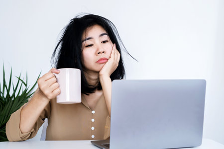 Asian businesswoman tired of working at office desk hand holding coffee mug looking at laptop lack of energyの写真素材