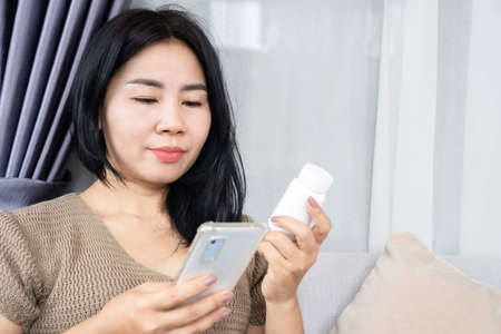 Asian woman reading a prescription medication label hand holding bottle of drug and checking information on mobile phoneの写真素材