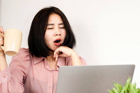 fatigued Asian woman yawning and clutching a cup of coffee while seated at her office deskの写真素材