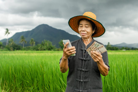 smiling Asian woman farmer holds Thai banknotes and a mobile phone, illustrating the benefits of...