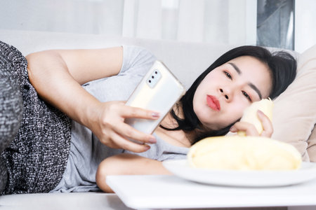relaxed Asian woman indulging in durian fruit while using phone, illustrating the concept of unhealthy lifestyleの写真素材