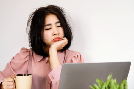 Asian woman Feeling tired or lacking energy after waking up and not feeling refreshed to start the day at the workplace hand holding a glass of coffee at the office deskの写真素材