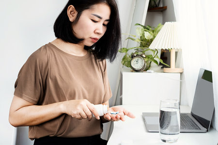 Asian woman taking some painkiller pills from a bottle while working on computer at deskの写真素材